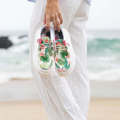 A woman walking on the beach with the ocean in the background with a pair of Gator Waders Camp Shoes in her hand.