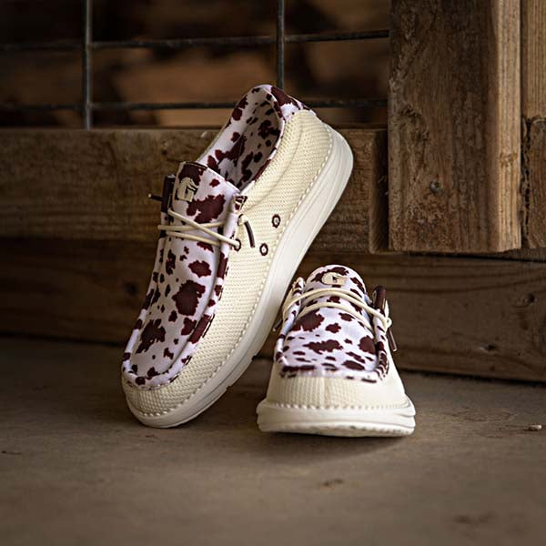 A pair of Camp Shoes in cow print sitting on the floor.