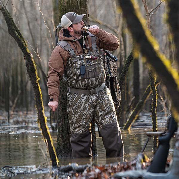 Man standing in water by a tree blowing a duck call